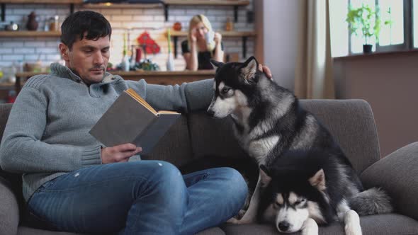 A Man Stroking a Husky Sitting on a Couch and Reading a Book. Woman Drinking Coffee Standing in the