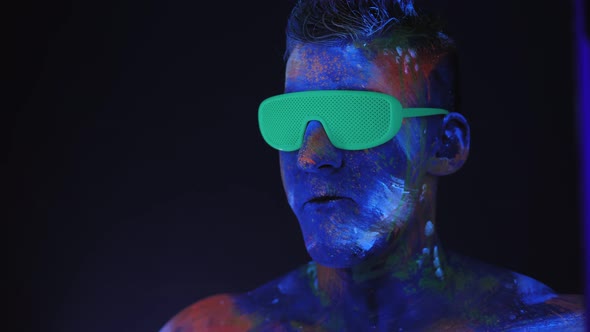 Man with UV Drawings on His Face and Neon Glasses Inflates a Bubble of Bubblegum