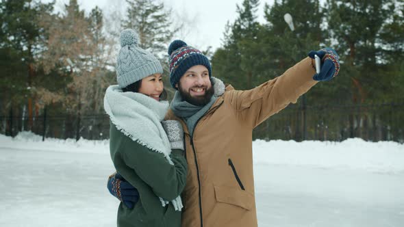 Slow Motion of Joyful Guy Taking Selfie with Asian Girlfriend at Ice Skating Rink Using Smartphone