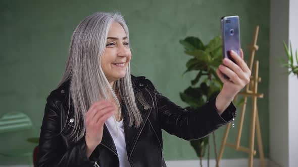 Joyful Senior Woman with Straight Gray Hair in Black Jacket Looking at Phone Camera at Home