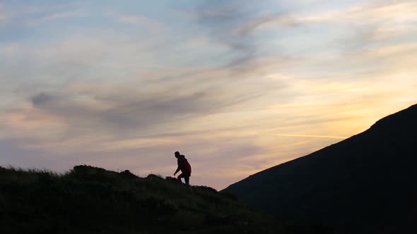 Dark silhouette of a hiker climbing a mountain at sunset and raising his hands reaching summit