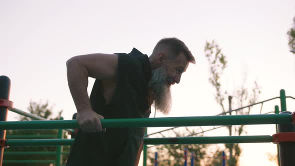 Fit Handsome Middle Aged Man with Long Gray Beard Doing Push Ups on Parallel Bars on Sports Ground