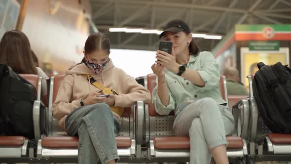 A Woman and Her Daughter with Smartphones in Their Hands are Sitting at the Airport in the Waiting