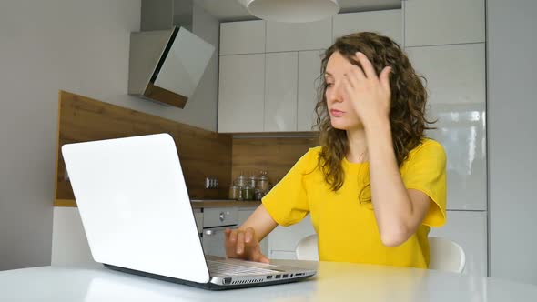 Young Woman with Curly Hair and Yellow Shirt is Working From Home Using Her Laptop at the Kitchen