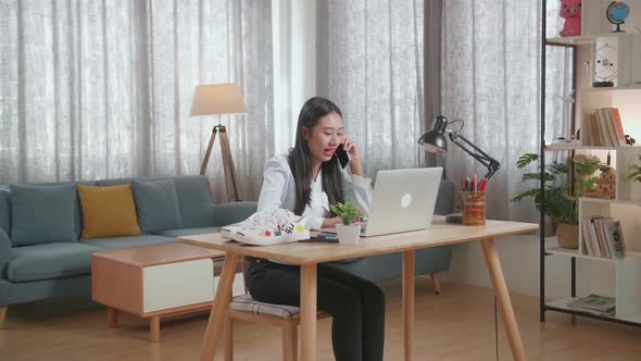 Asian Female Footwear Designer Talking On Smartphone While Working On A Laptop At Home