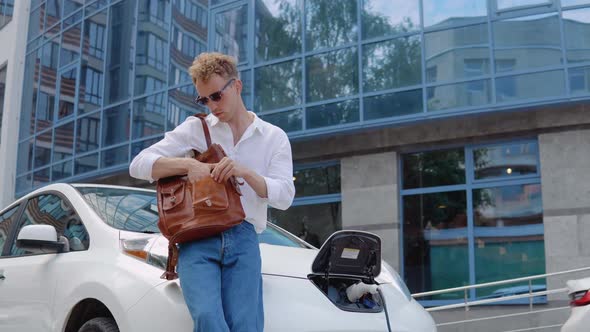 Stylish Modern Young Curly Man with a Stylish City Briefcase Walks Away From His Electric Car