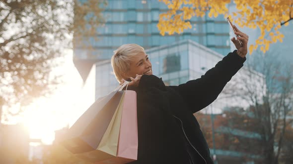 Young Woman Shopaholic Standing in the Park with Shopping Bags and Using Smartphone To Take Selfie