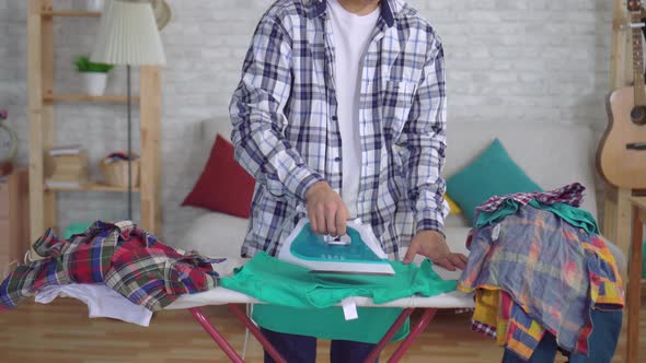 Young Asian Man Ironing Clothes on Ironing Board Looking at the Camera and Smiling