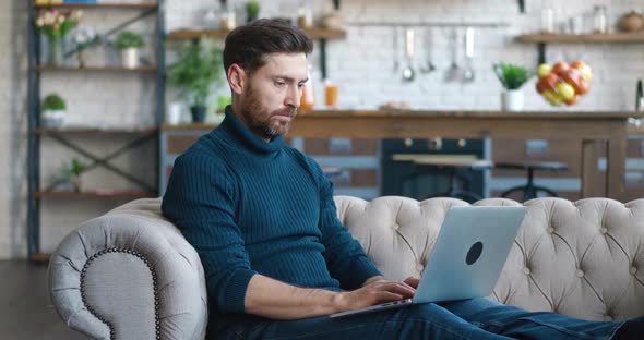 Handsome Millennial Man Working on Laptop Computer While Sitting on a Sofa in Cozy Living Room