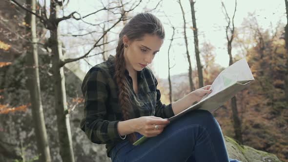 Girl with Long Hair Braided in a Braid, Sitting on a Rock in the Woods and Looking at the Map