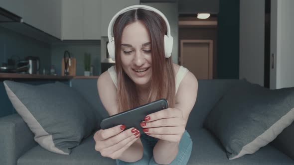 Millennial Female with Headphones Sitting with Plays a Smartphone