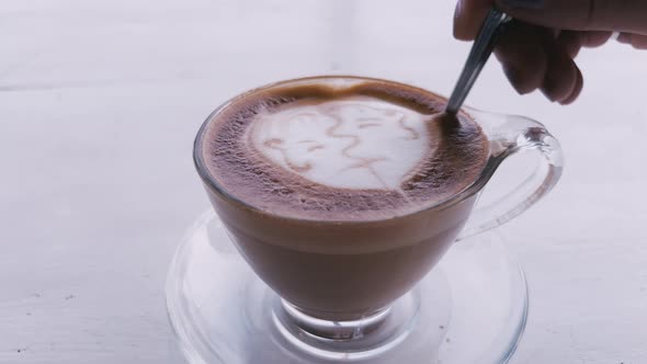 A Transparent Coffee Mug with a Latte Art Foam Pattern in Form of a Kissing Couple Stands on a