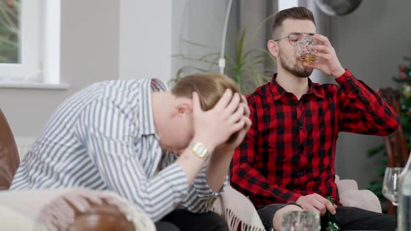 Young Man in Eyeglasses Drinking Scotch and Looking at Empty Beer Bottle with Blurred Frustrated