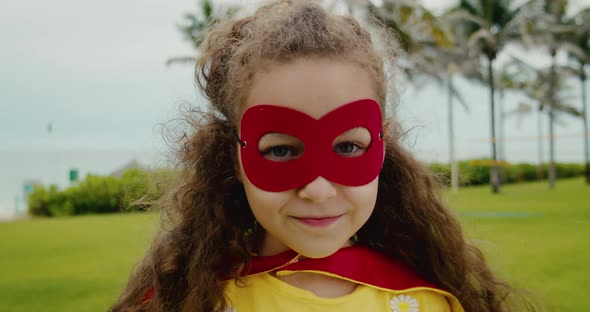 Portrait of a Cute Funny Child Girl in a Superhero Costume in a Red Cloak and a Red Mask Playing a