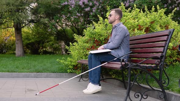 Blind Man Reading Braille Book Sitting on Bench in Summer Park