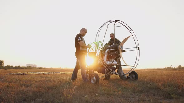 Motor Paraglider Stands in a Field at Sunset with a Wooden Propeller, Two Pilots Warm Up the Engine