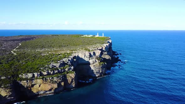 Drone flying high over blue ocean showing huge sea side cliffs. A lighthouse can be seen in the back