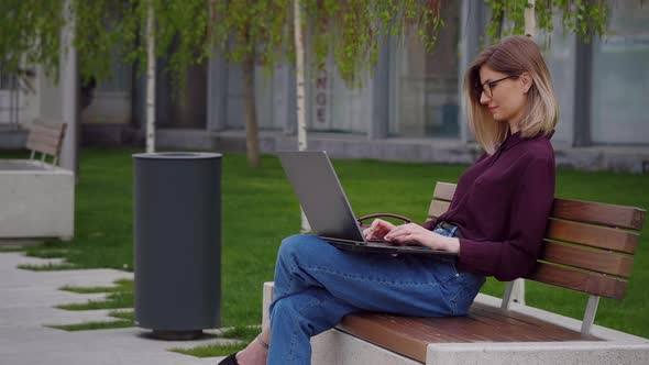 Young pretty business woman works on laptop outdoor sitting on park bench.