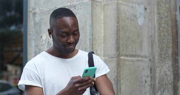 Close Up Shot of Handsome African American Guy in Casual Clothes with Earphones Using His Smartphone