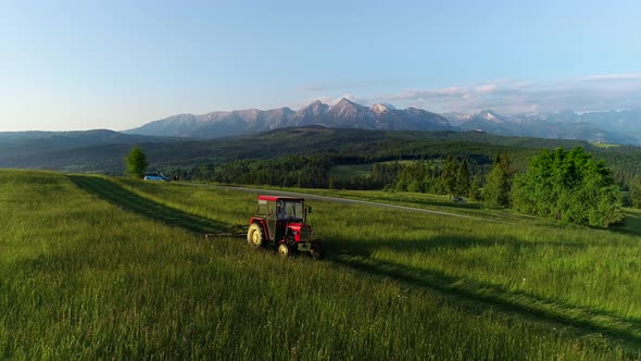 Tractor mowing the grass with beautiful high mountains in the background, aerial view.