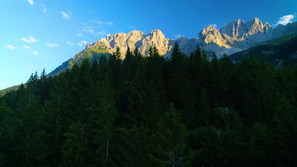 Epic aerial view ascent over woodland towards sunlit golden Wilder Kaiser mountains peaks