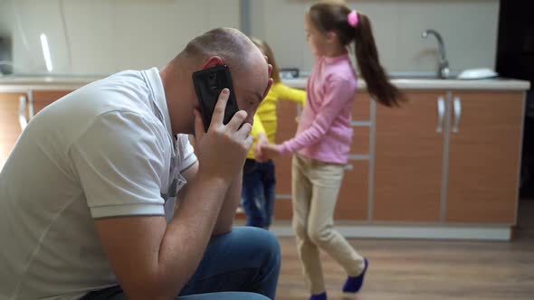 Tired Father Speaking on Smartphone Near Playing Daughters