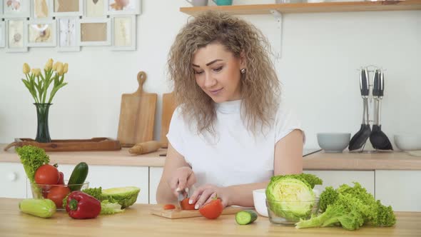 Young Blonde Woman with Curly Hair Slices a Red Tomato Sitting at a Table in the Kitchen in a