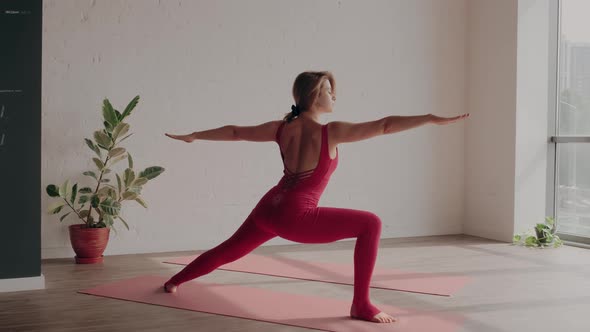 Woman in Red Sports Uniform Stands in Warrior Pose in Yoga Studio