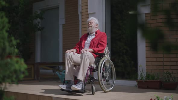 Wide Shot of Old Disabled Caucasian Man in Wheelchair Enjoying Sunny Summer Day Outdoors on House