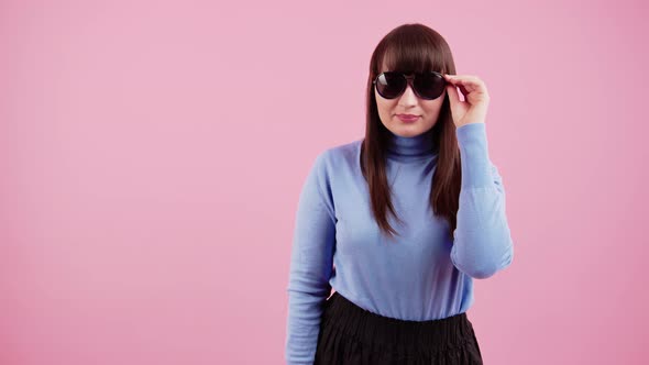 Portrait of a Girl in a Blue Turtleneck on a Pink Background Who Takes Off Her Sunglasses and Smiles