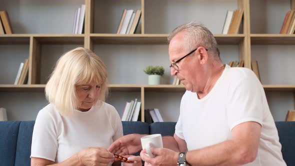 An Elderly Couple of European Age is Sitting on the Sofa the Husband is Giving His Wife Pills