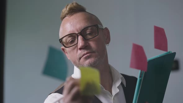 Intelligent Focused Caucasian Man in Eyeglasses with Mohawk Haircut Writing on Sticky Notes on Glass