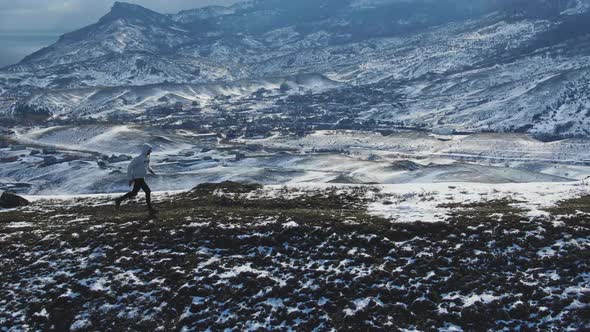 An Athlete Jogs Along a Snowcovered Trail at the Top of a Hill Against the Backdrop of a Mountain