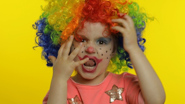 Little Child Girl Clown in Colorful Wig Hides Behind Her Hands and Shows Funny Faces. Halloween