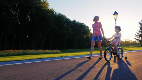 Mother and Daughter Walking in the Park. Mother Holds Her Hand, a Child Riding a Bicycle Near