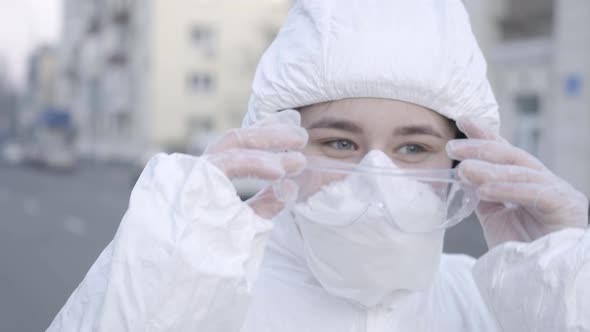 Close-up Face of Smiling Young Woman in Face Mask and Antiviral Suit Putting on Protective