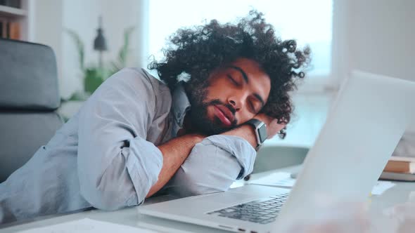 Young African American Man Freelancer Sleeps with Head on Table Next to Laptop