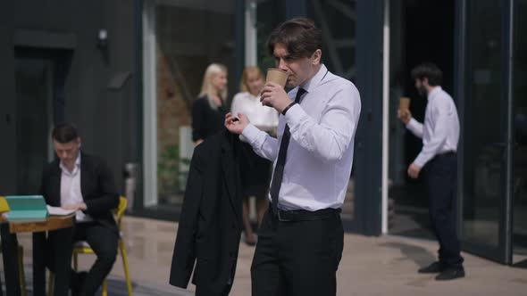 Confident Positive Businessman Drinking Coffee Outdoors on Terrace Thinking with Blurred Colleagues