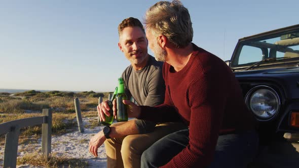 Happy caucasian gay male couple sitting by car drinking bottles of beer on sunny day at the beach