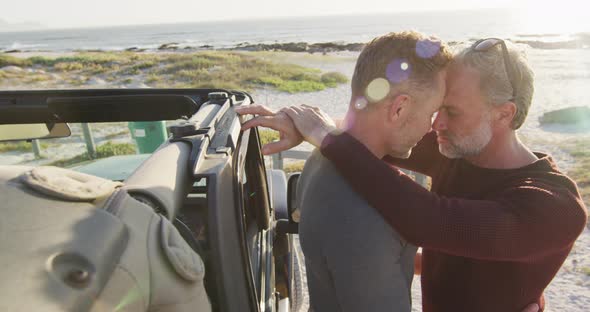 Happy caucasian gay male couple standing by car embracing on sunny day at the beach