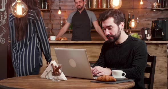 A Caucasian Man with Beard Works at His Laptop While Customers Enter the Coffee Shop