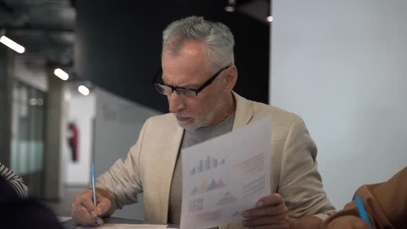 Serious senior businessman wearing glasses sitting at the table with his team