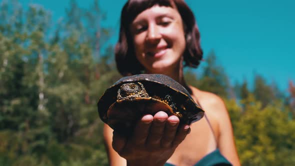 Woman Holds Funny Turtle in Arm and Smiles on River with Green Vegetation