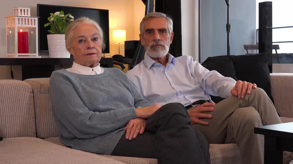 An Elderly Couple Sits on A Couch in An Apartment and Looks Seriously at The Camera