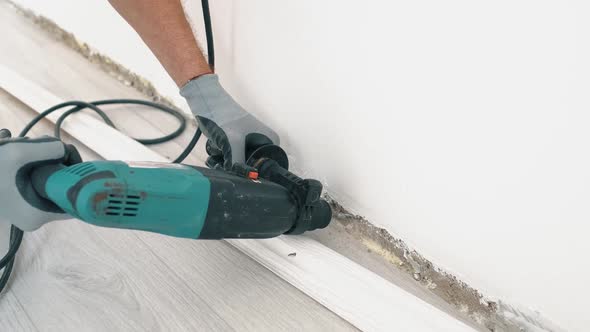 A Man in Work Gloves Makes a Hole with a Puncher in the Wall Screwing the Skirting Board to the Wall