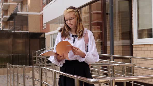 A Teenage Girl with Glasses is Standing in the School Yard with an Open Notebook