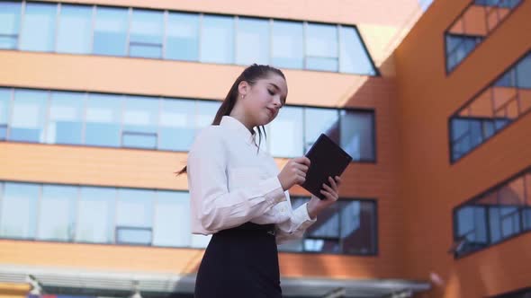 Young Business Girl Stands Near Business Building Holding a Tablet in Hands