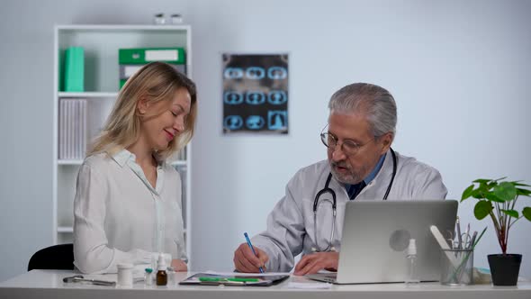 Man Doctor in White Medical Coat Sits at Table in Clinic Looks at Snapshot of Patient and Speaking