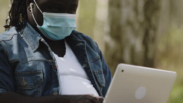 Remote Work Concept. African Man with Medical Mask Using Laptop in the Park