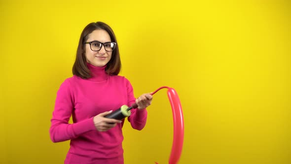 A Young Woman Inflates a Long Red Balloon with a Pump on a Yellow Background. Girl in a Pink